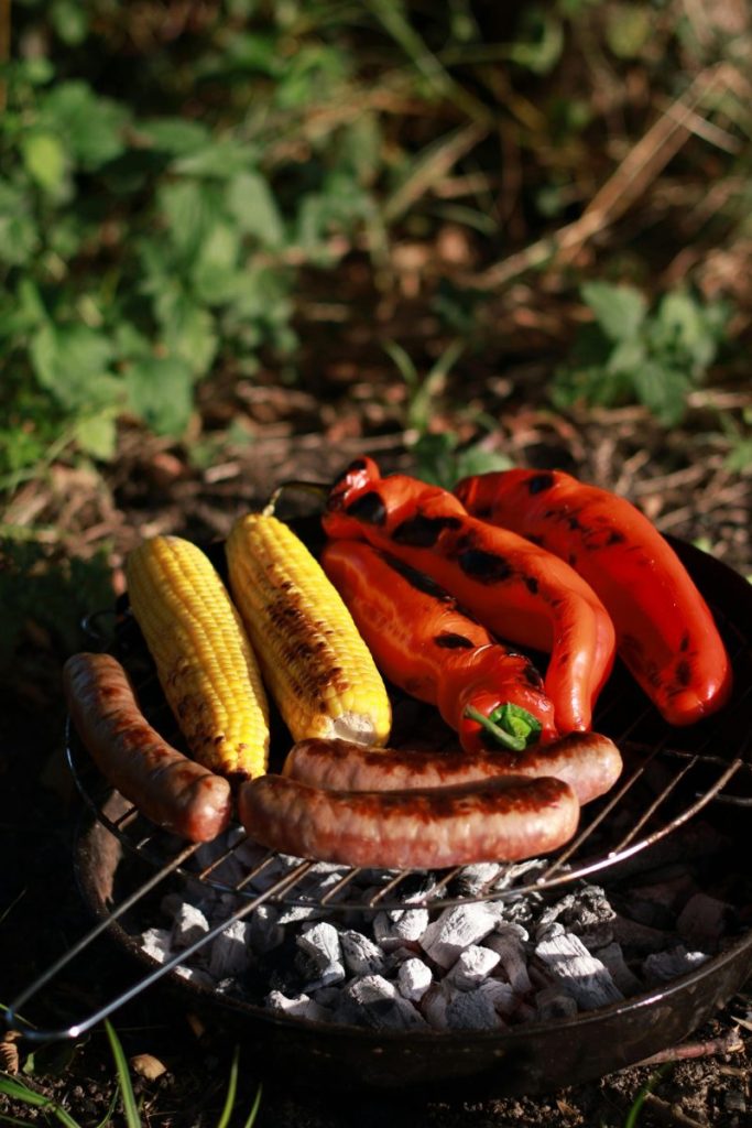 Grilled sausages, corn, and red peppers on a barbecue outdoors.